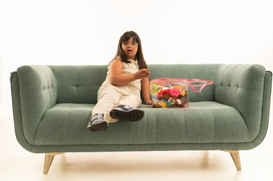 الانتباه الى شئ ما بعيد , بنت صغيرة عربة خليجية سعودية تعاني من متلازمة داون ترتدي الزي البيج الكاجوال , صندوق الالعاب على اريكة باللون الرمادي , الجسم بالطول الكامل , بورتريه خلفية بيضاء