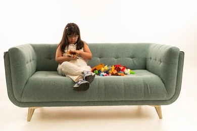 لعبة تنمية المهارات الذهنية المفيدة لاصحاب الاحتياجات الخاصة , بنت صغيرة عربية خليجية سعودية تعاني من متلازمة داون تحمل لعبة البازل , الجلوس على اريكة لون الرمادي , الجسم بالطول الكامل , بورتريه خلفية بيضاء