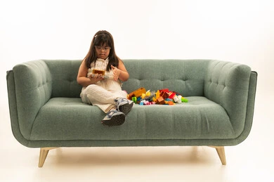 الانشغال في العاب الفك و التركيب المفيدة لاصحاب متلازمة داون , بنت صغيرة عربية خليجية سعودية من ذوي الهمم ترتدي الزي الكاجوال , الجلوس على الاريكة الرمادية بجانب العاب تنمية المهارات, الجسم بالطول الكامل , بورتريه خلفية بيضاء