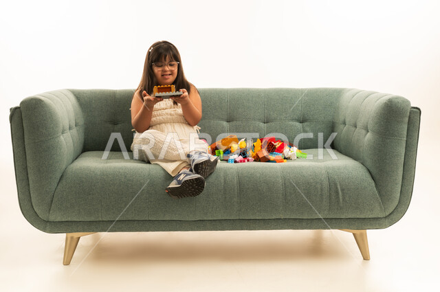 Expressions of happiness and joy after completing the fun puzzle, a small Arab Gulf Saudi girl with Down syndrome sitting on the gray sofa, full-body, portrait with a white background.