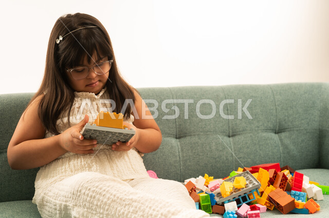 A small Arab Gulf Saudi girl with special needs wearing casual attire, holding a puzzle cube toy, sitting on a gray sofa, close-up portrait with a white background.