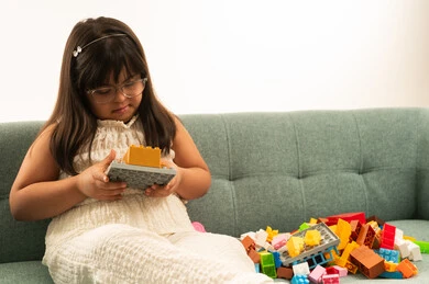 A small Arab Gulf Saudi girl with special needs wearing casual attire, holding a puzzle cube toy, sitting on a gray sofa, close-up portrait with a white background.