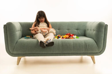 اللعب بالدمية الصغيرة , بنت صغيرة عربية خليجية سعودية من ذوي الهمم تجلس على الاريكة الرمادية , الجسم بالطول الكامل , ارتداء الزي الكاجوال , بورتريه خلفة بيضاء