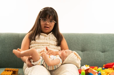 الجلوس على الاريكة باللون الرمادي , بنت صغيرة عربية خليجية سعودية من ذوي الهمم ترتدي الزي الكاجوال , اللعب بالدمية الصغيرة , النظر للكاميرا, بورتريه خلفية بيضاء , الجسم بالطول الكامل