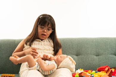 بنت صغيرة من اصحاب متلازمة داون تلعب بالدمية الصغيرة , الجلوس على اريكة رمادية اللون , العاب فك و تركيب خاصة بتطوير المهارات الذهنية , الجسم بالطول الكامل , بورتريه خلفية بيضاء