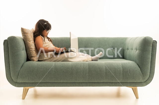 وضع جهاز الحاسب المتنقل على الارجل الممدودة , بنت عربية خليجية سعودية صغيرة من اصحاب متلازمة داون تنظر الى شاشة الحاسوب , الجسم بالطول الكامل , بورتريه خلفية بيضاء