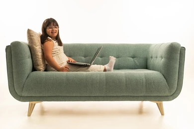 النظر مع الابتسامة للكاميرا , بنت صغيرة عربية خليجية سعودية من ذوي الهمم تجلس على الاريكة الرمادية اللون ممدة رجلها , بورتريه خلفية بيضاء, استخدام جهلز الحاسب المتنقل , الجسم بالطول الكامل