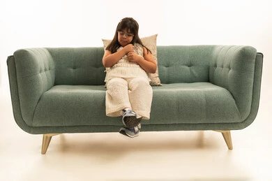 بنت صغيرة عربية خليجية سعودية من اصحاب متلازمة داون تمسك شعرها في حنان , الجلوس على الاريكة الرمادية اللون , الجسم باطول الكامل , بورتريه خلفية بيضاء