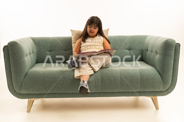 Reading aloud, a Saudi Gulf Arab girl with special needs wearing casual attire, sitting on a gray sofa while reading, portrait with a white background, full body.