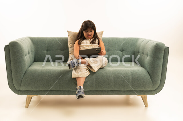 استخدام التطبيقات التقنية الحديثة الخاصة باصحاب متلازمة داون , بنت صغيرة عربية خليجية سعودية من ذوي الهمم تحمل الجهاز اللوحي , الجلوس على اريكة رمادية اللون , بورتريه الجسم بالطول الكامل , خلفية بيضاء