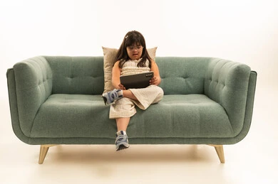 استخدام التطبيقات التقنية الحديثة الخاصة باصحاب متلازمة داون , بنت صغيرة عربية خليجية سعودية من ذوي الهمم تحمل الجهاز اللوحي , الجلوس على اريكة رمادية اللون , بورتريه الجسم بالطول الكامل , خلفية بيضاء