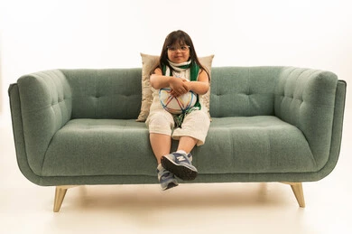 الامساك بكرة القدم بين اليدين , بنت صغيرة من ذوي الهمم عربية خليجية سعودية ترتدي الز الكاجوال تجلس على اريكة رمادية اللون , الجسم بالطول الكامل , بورتريه خلفية بيضاء , النظر و ايماءات الابتسامة للكاميرا