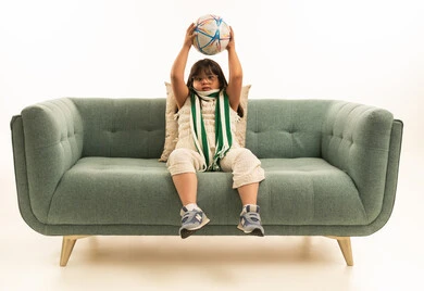 العاب تناسب ذوي الاحتياجات الخاصة, بنت عربية خليجية سعودية من ذوي الهمم ترتدي الزي الكاجوال تجلس على اريكة رمادية اللون , الجسم بالطول الكامل , رفع كرة القدم للاعلى , بورتريه خلفية بيضاء