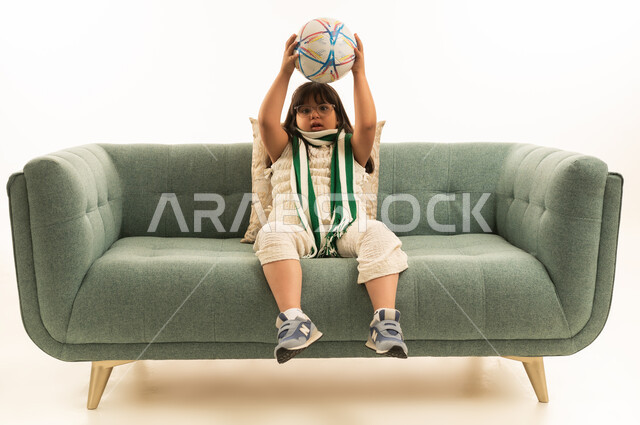 A small Arab Gulf Saudi girl with special needs wearing casual attire raises a soccer ball above her head, looking at the camera, wearing a green keffiyeh, sitting on a gray sofa, with Down syndrome, full body, white background.