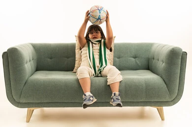 A small Arab Gulf Saudi girl with special needs wearing casual attire raises a soccer ball above her head, looking at the camera, wearing a green keffiyeh, sitting on a gray sofa, with Down syndrome, full body, white background.