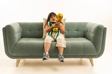 النظر الى الكأس الذهبية , بنت صغيرة عربية خليجية سعودية ترتدي الزي الكاجوال تجلس على الاريكة الرمادية , وضع الكوفية الخضراء حول الرقبة , الالعاب البارالمبية لذوي الاحتياجات الخاصة , الجسم بالطول الكامل , بورتريه خلفية بيضاء