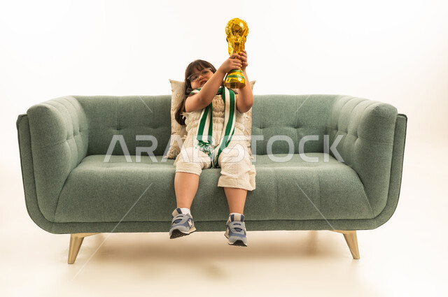 رفع الكأس الذهبية بعد الفوز في الالعاب البارالمبية , بنت صغيرة عربية خليجية سعودية ترتدي الزي الكاجوال تجلس على الاريكة الرمادية , ارتداء ميداليات النصر , بورتريه الجسم بالطول الكامل , خلفية بيضاء