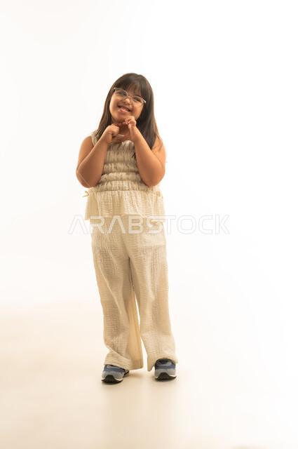 The sign with a heart symbol, a small girl with determination, an Arab Gulf Saudi, wearing casual attire, looking at something, full body, portrait with a white background, smiling at the camera.