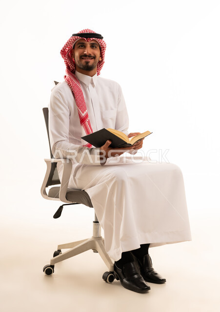 رجل سعودي بالزي التقليدي يجلس على كرسي مكتب يحمل كتابا