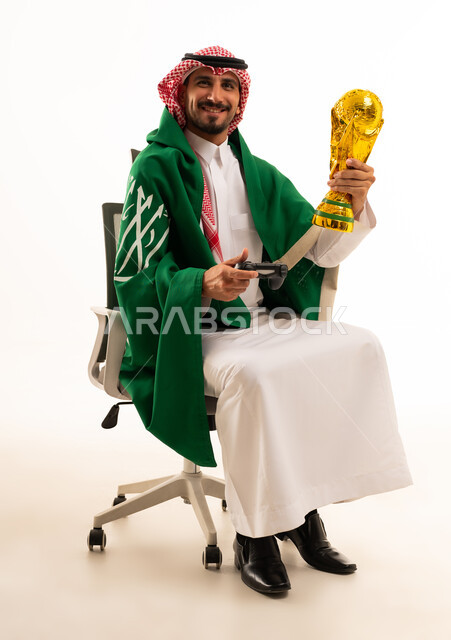 شاب سعودي بالزي التقليدي يحمل كأس ذهبية وجهاز ألعاب