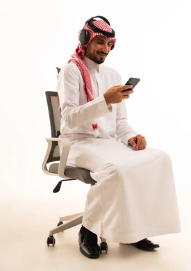 رجل سعودي يستخدم الهاتف الذكي والسماعات على كرسي مكتب