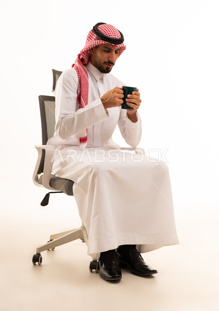 رجل سعودي بالزي التقليدي يمسك كوباً على كرسي مكتب