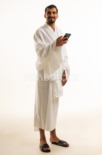 رجل سعودي بملابس الإحرام يمسك هاتفاً ذكياً بخلفية بيضاء