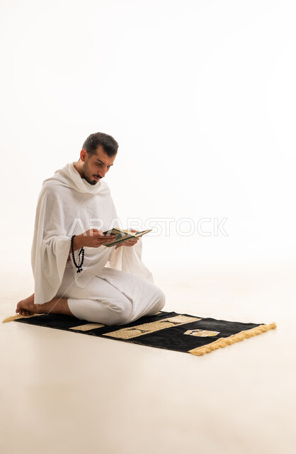 رجل مسلم بملابس الإحرام يقرأ القرآن على سجادة الصلاة