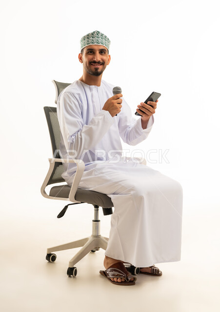 رجل عماني يحمل ميكروفون وهاتف ذكي على كرسي مكتب