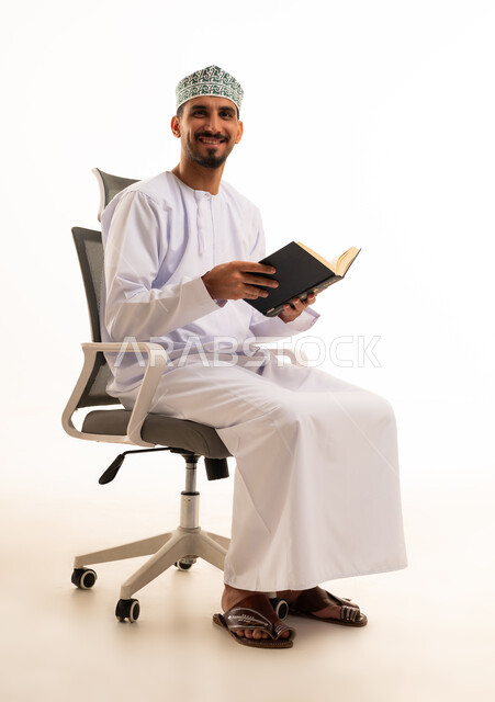 رجل عماني مبتسم يحمل كتاباً على كرسي مكتب