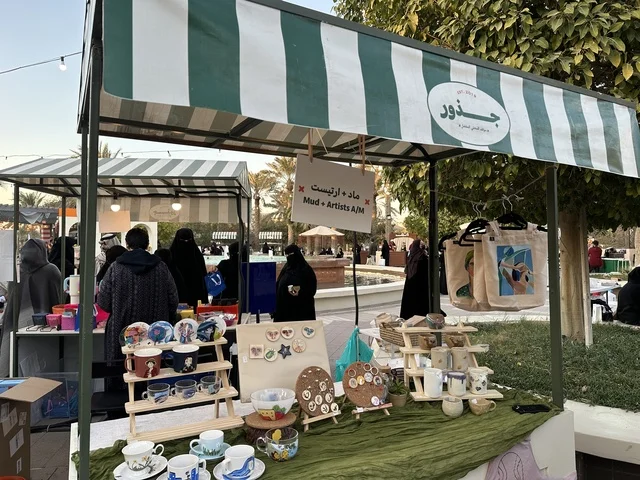 أركان تسوق خارجية في سوق جذور في حديقة الأمير سعود بمدينة الظهران، المنطقة الشرقية، المملكة العربية السعودية، سوق مجتمعي مفتوح يدعم المشاريع المحلية، منتجات حرفية وأطعمة منزلية، فعاليات عائلية وأنشطة ترفيهية، أجواء تراثية واجتماعية نابضة بالحياة