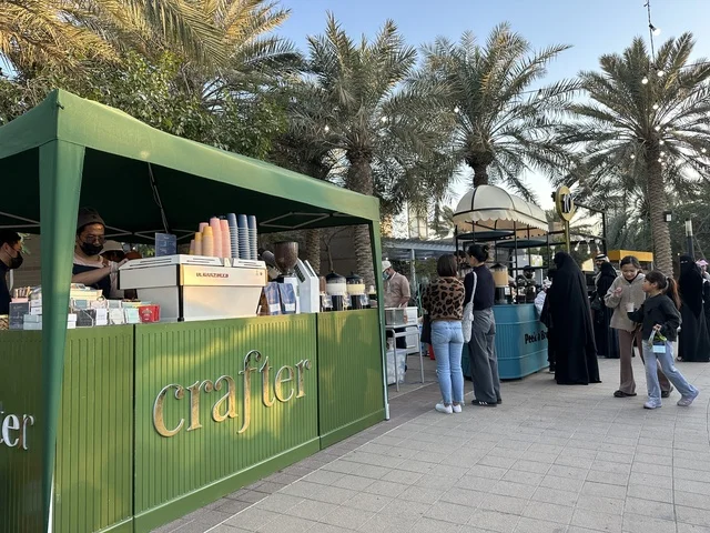 Visitors gathered at the Jood Market in Prince Saud Park in Dhahran, Eastern Province, Saudi Arabia. It is an open community market that supports local projects, featuring artisanal products and homemade foods, outdoor shopping areas, family events, and recreational activities, all set in a vibrant heritage and social atmosphere.