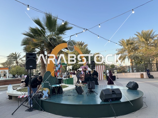A live music performance at the Jood Market in Prince Saud Park in Dhahran, Eastern Province, Saudi Arabia, an open community market that supports local projects, artisanal products, and homemade foods, outdoor shopping corners, family events, and recreational activities, with a vibrant heritage and social atmosphere.