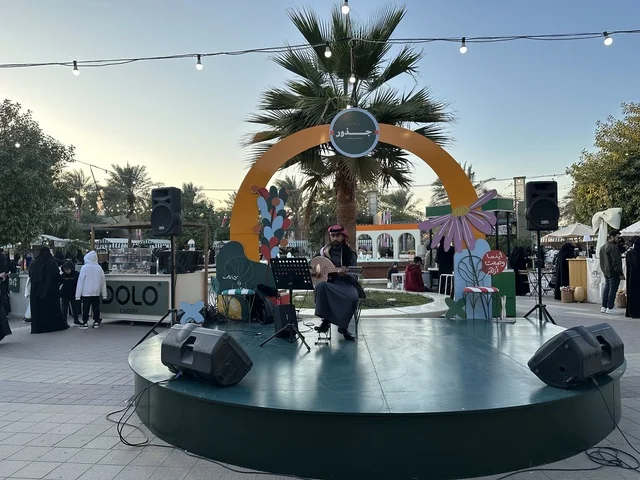 A live music performance at the Jood Market in Prince Saud Park in Dhahran, Eastern Province, Saudi Arabia, an open community market that supports local projects, artisanal products, and homemade foods, outdoor shopping corners, family events, and recreational activities, with a vibrant heritage and social atmosphere.