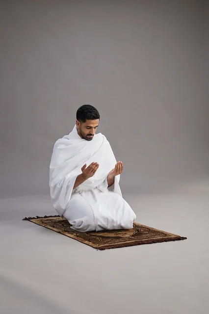 بورتريه لرجل عربي سعودي خليجي بملابس الإحرام يدعو رافعاً يديه على سجادة الصلاة, اداء مناسك الحج والعمره,  خلفيه رماديه