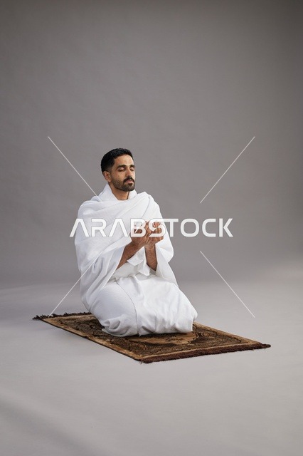 بورتريه لرجل عربي سعودي خليجي بملابس الإحرام يدعو رافعاً يديه على سجادة الصلاة, اداء مناسك الحج والعمره,  خلفيه رماديه