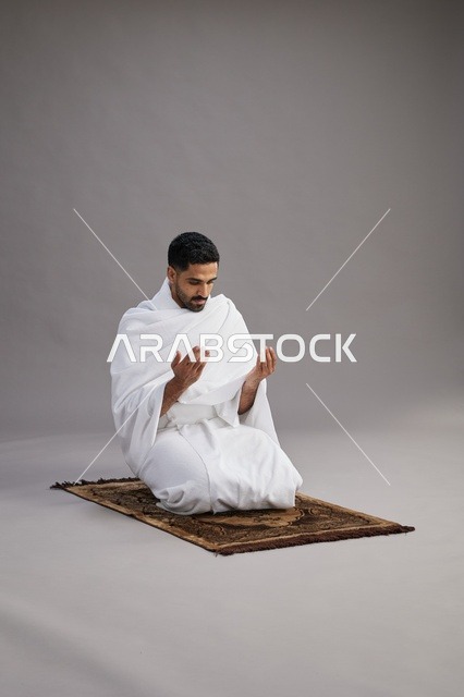 بورتريه لرجل عربي سعودي خليجي بملابس الإحرام يدعو رافعاً يديه على سجادة الصلاة, اداء مناسك الحج والعمره,  خلفيه رماديه