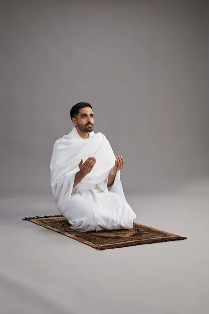 A portrait of an Arab Saudi Gulf man in Ihram clothing, praying with his hands raised on a prayer rug, performing the rituals of Hajj and Umrah, with a gray background.