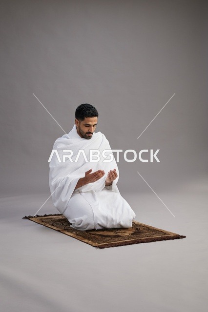 A portrait of an Arab Saudi Gulf man in Ihram clothing, praying with his hands raised on a prayer rug, performing the rituals of Hajj and Umrah, with a gray background.