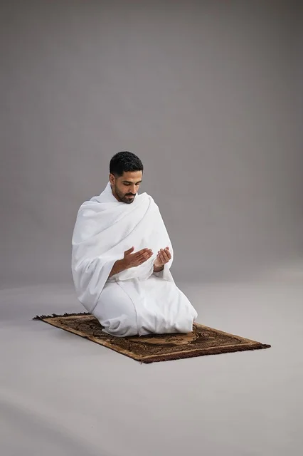 A portrait of an Arab Saudi Gulf man in Ihram clothing, praying with his hands raised on a prayer rug, performing the rituals of Hajj and Umrah, with a gray background.