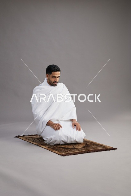 A portrait of an Arab Saudi Gulf man in Ihram clothing praying on a prayer rug, performing the rituals of Hajj and Umrah, with a gray background.
