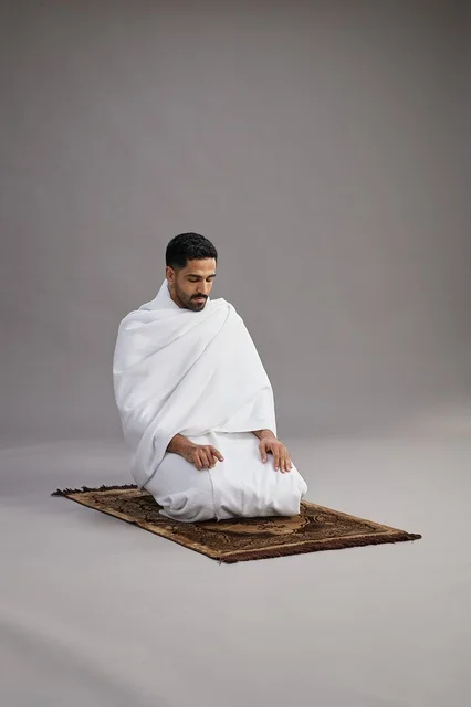 بورتريه لرجل عربي سعودي خليجي بملابس الإحرام يصلي على سجادة الصلاة, اداء مناسك الحج والعمره,  خلفيه رماديه