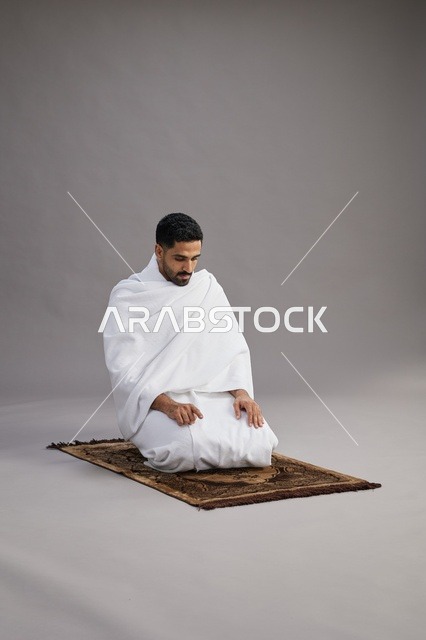 بورتريه لرجل عربي سعودي خليجي بملابس الإحرام يصلي على سجادة الصلاة, اداء مناسك الحج والعمره,  خلفيه رماديه
