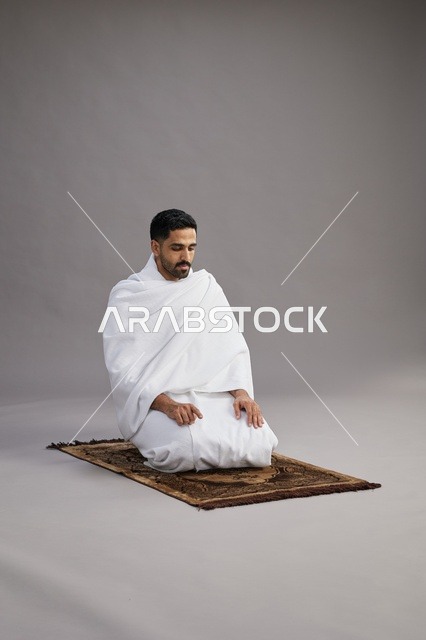 بورتريه لرجل عربي سعودي خليجي بملابس الإحرام يصلي على سجادة الصلاة, اداء مناسك الحج والعمره,  خلفيه رماديه