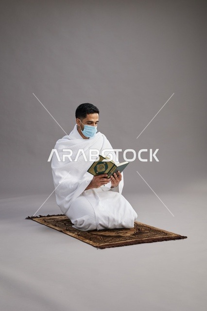 بورتريه لرجل عربي سعودي خليجي بملابس الإحرام يرتدي الكمامه ويقرا القران على سجادة الصلاة, اداء مناسك الحج والعمره,  خلفيه رماديه