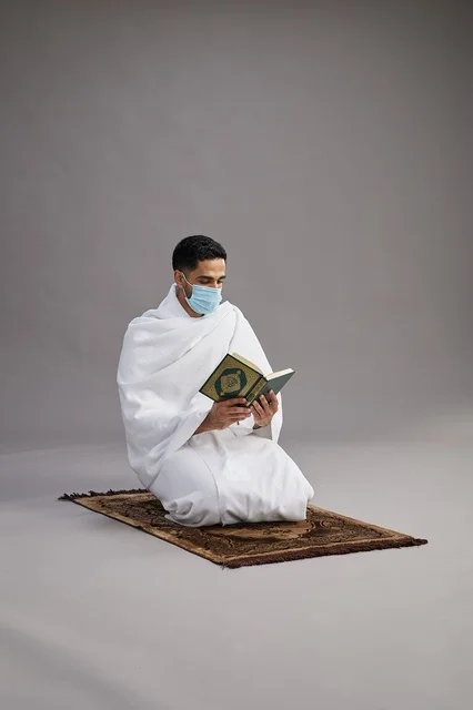 بورتريه لرجل عربي سعودي خليجي بملابس الإحرام يرتدي الكمامه ويقرا القران على سجادة الصلاة, اداء مناسك الحج والعمره,  خلفيه رماديه
