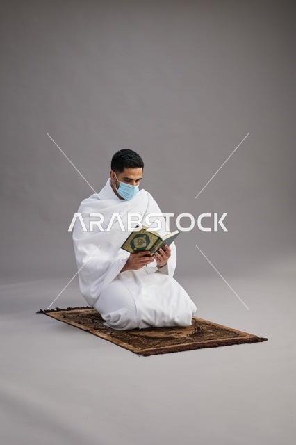 بورتريه لرجل عربي سعودي خليجي بملابس الإحرام يرتدي الكمامه ويقرا القران على سجادة الصلاة, اداء مناسك الحج والعمره,  خلفيه رماديه