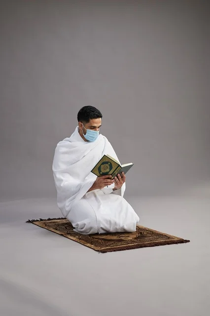 A portrait of an Arab Saudi Gulf man in Ihram clothing wearing a mask and reading the Quran on a prayer rug, performing the rituals of Hajj and Umrah, with a gray background.