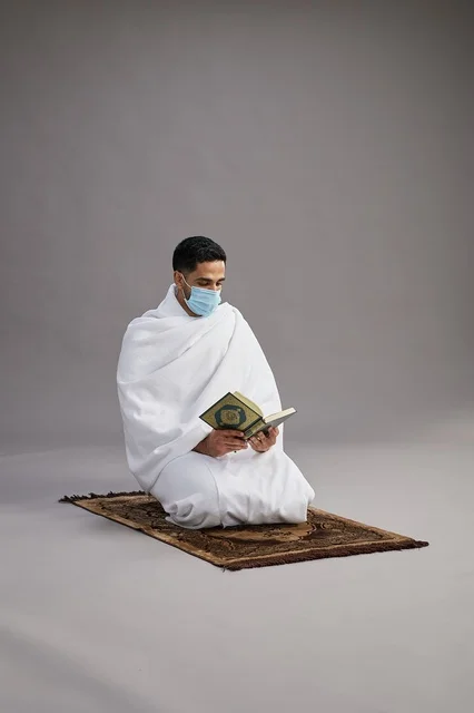 بورتريه لرجل عربي سعودي خليجي بملابس الإحرام يرتدي الكمامه ويقرا القران على سجادة الصلاة, اداء مناسك الحج والعمره,  خلفيه رماديه