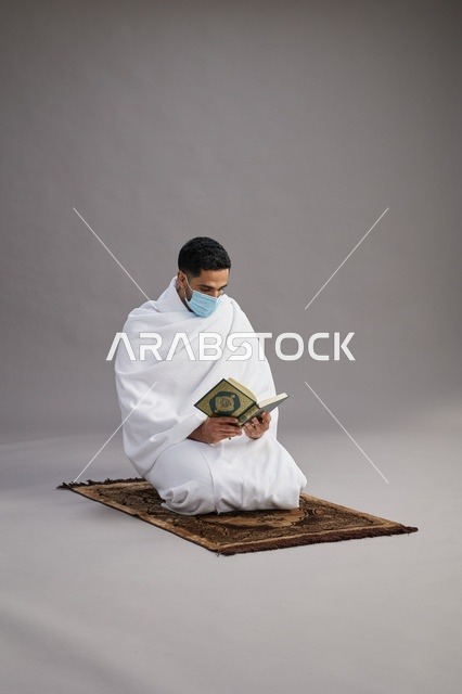 بورتريه لرجل عربي سعودي خليجي بملابس الإحرام يرتدي الكمامه ويقرا القران على سجادة الصلاة, اداء مناسك الحج والعمره,  خلفيه رماديه